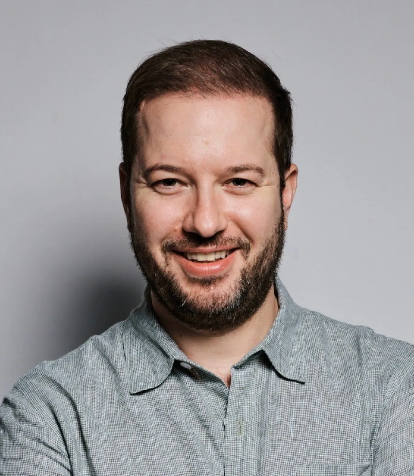 Smiling man with short dark hair and beard wearing a light gray collared shirt against a plain gray background.