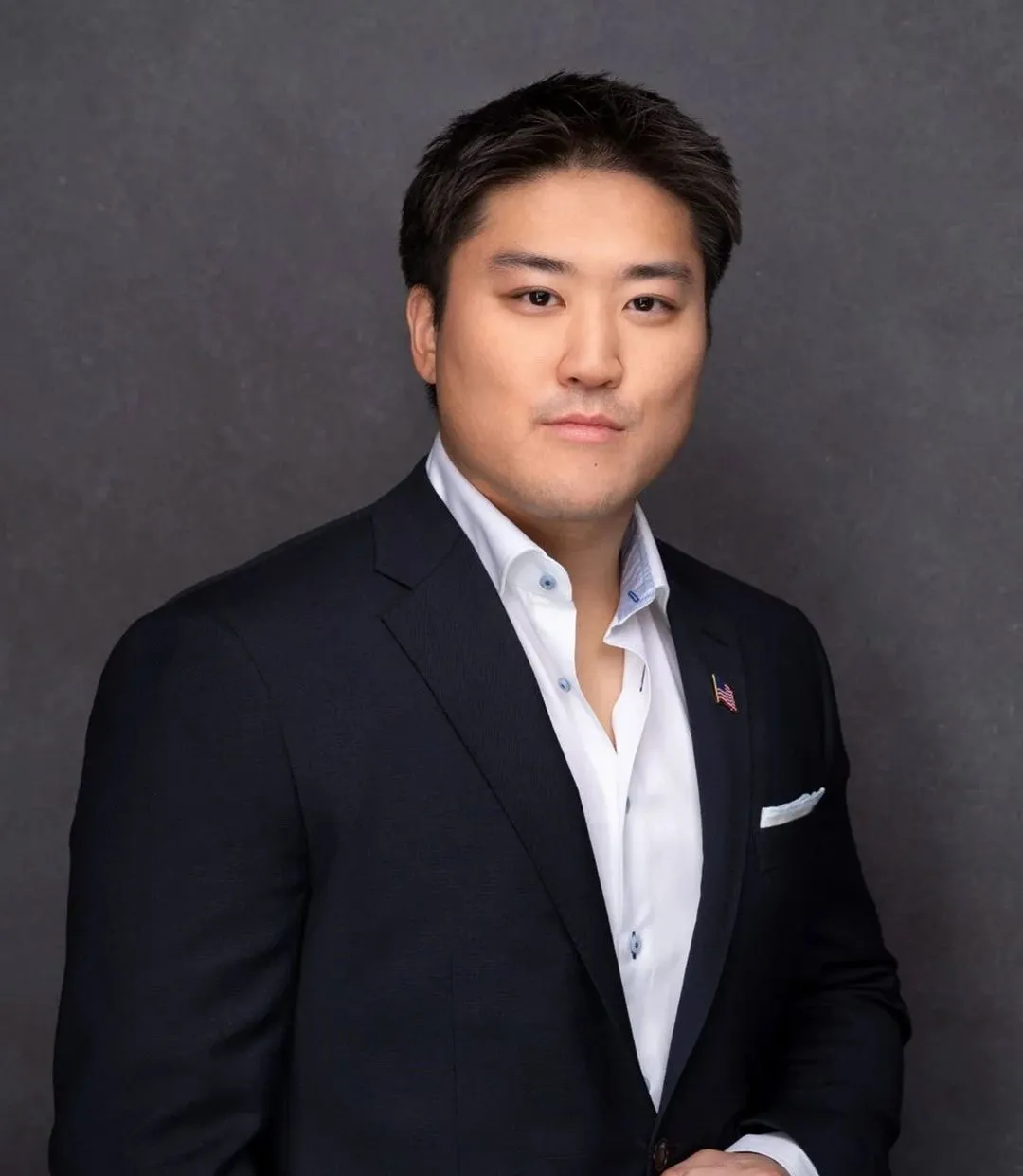 Man wearing a dark blazer with a small American flag pin, white dress shirt, and pocket square, posing against a gray background.