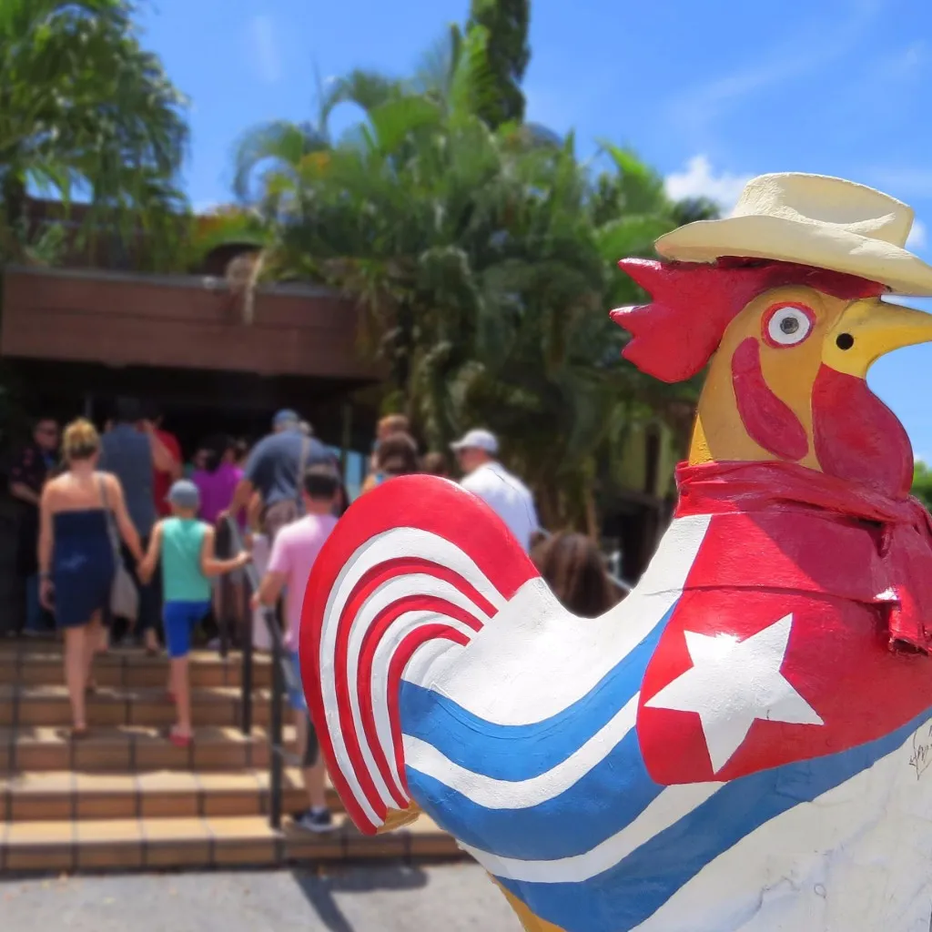 Colorful rooster statue wearing a hat and scarf painted with the Cuban flag in a sunny outdoor setting with people walking up stairs in the background.