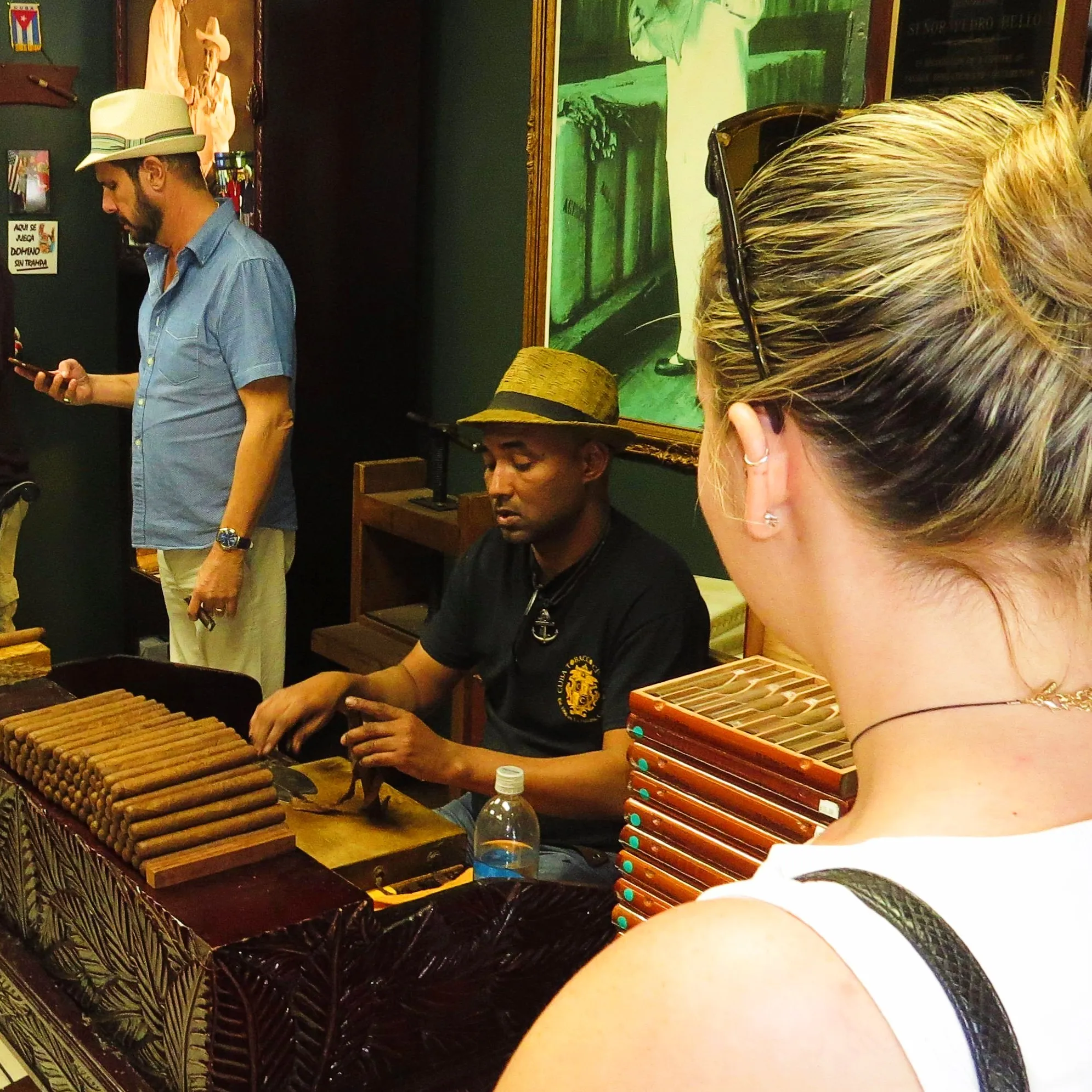 Man in a straw hat rolling cigars by hand at a wooden counter with cigar boxes, observed by a woman with sunglasses on her head and a man in a blue shirt using a phone in the background.