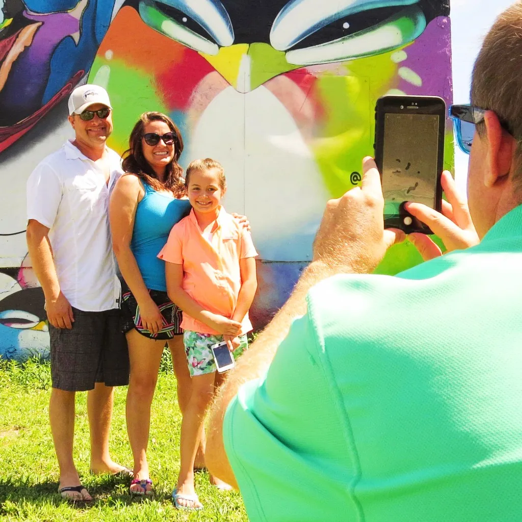 Person in green shirt taking photo of smiling family of three standing on grass in front of colorful graffiti wall.