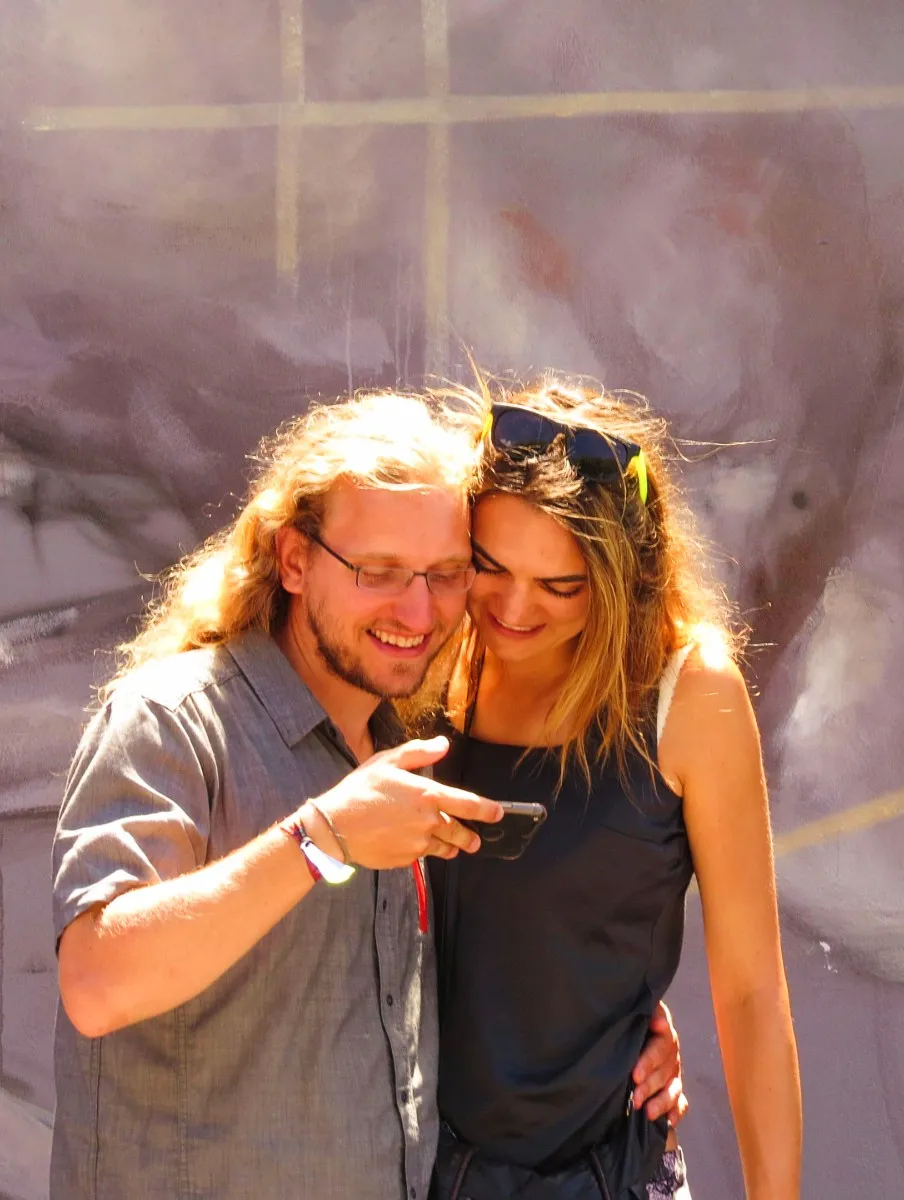 Smiling man and woman standing close together outdoors, looking at a smartphone.