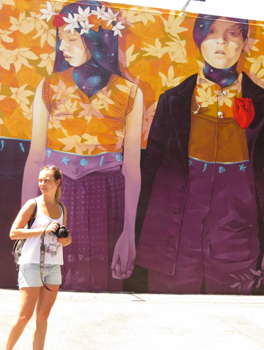 Woman holding a camera standing in front of a colorful mural depicting two people holding hands with floral and cosmic designs.