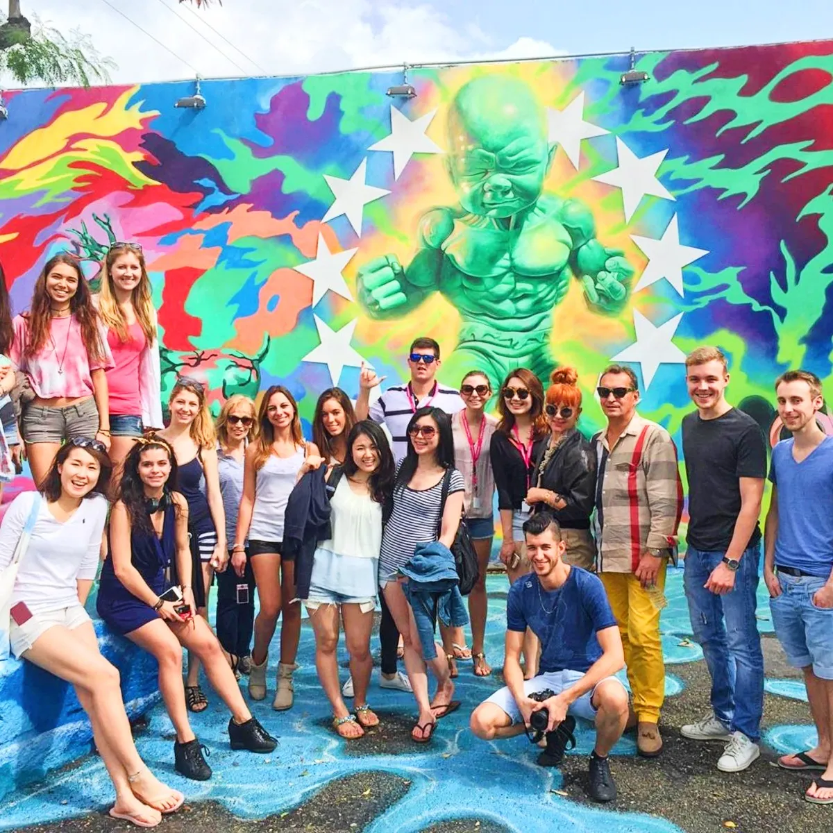 Group of diverse young adults posing cheerfully in front of a colorful mural featuring a muscular green baby figure surrounded by white stars.