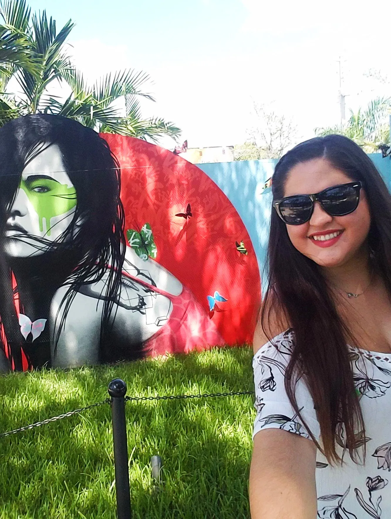 Smiling woman with long dark hair and sunglasses taking a selfie in front of a colorful mural of a woman with green face paint and butterflies on a sunny day.