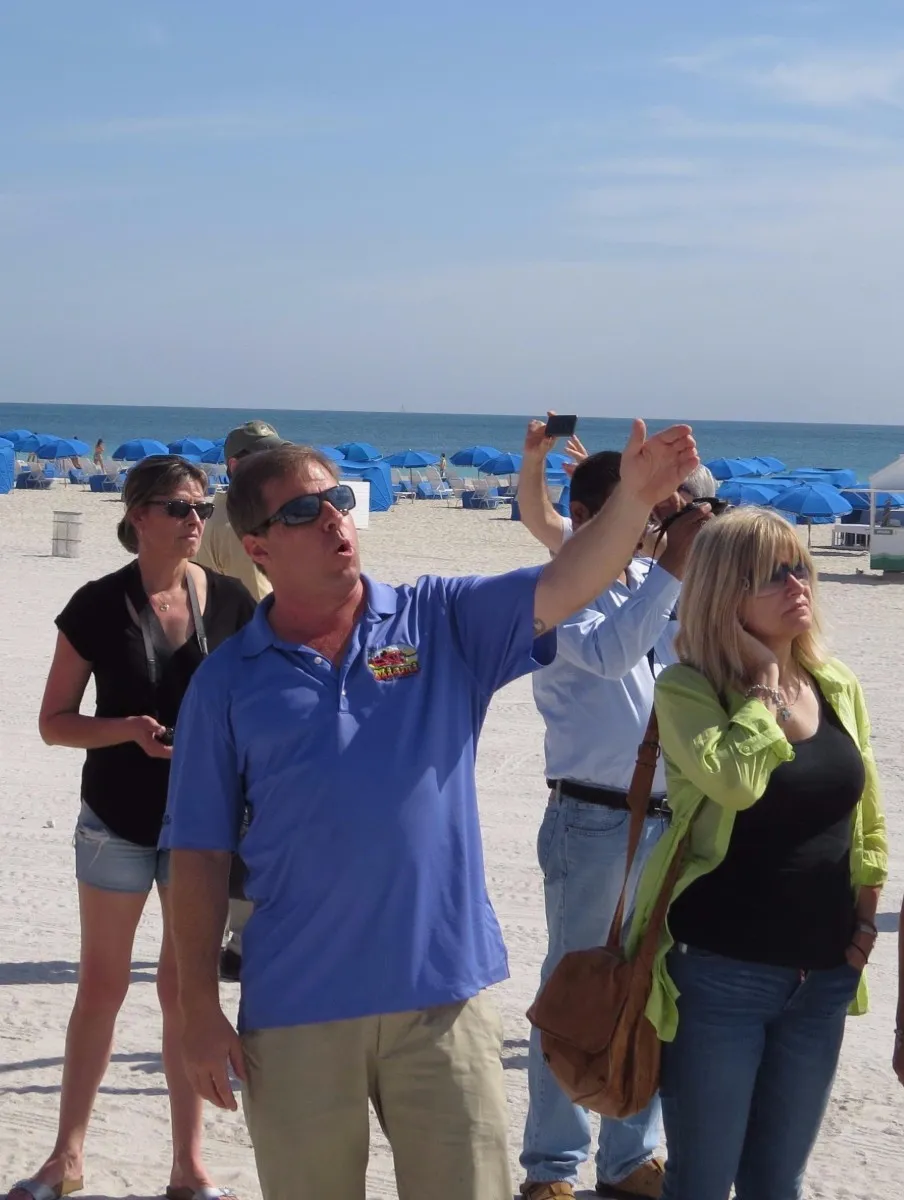 Group of people on a sunny beach with blue umbrellas in the background, one man in a blue shirt gestures while others listen or take photos.