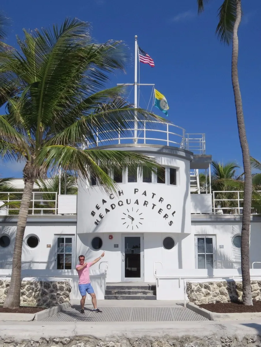 Man in pink shirt and blue shorts pointing at a white Beach Patrol Headquarters building with American and yellow-green flags flying atop, surrounded by palm trees under a clear blue sky.