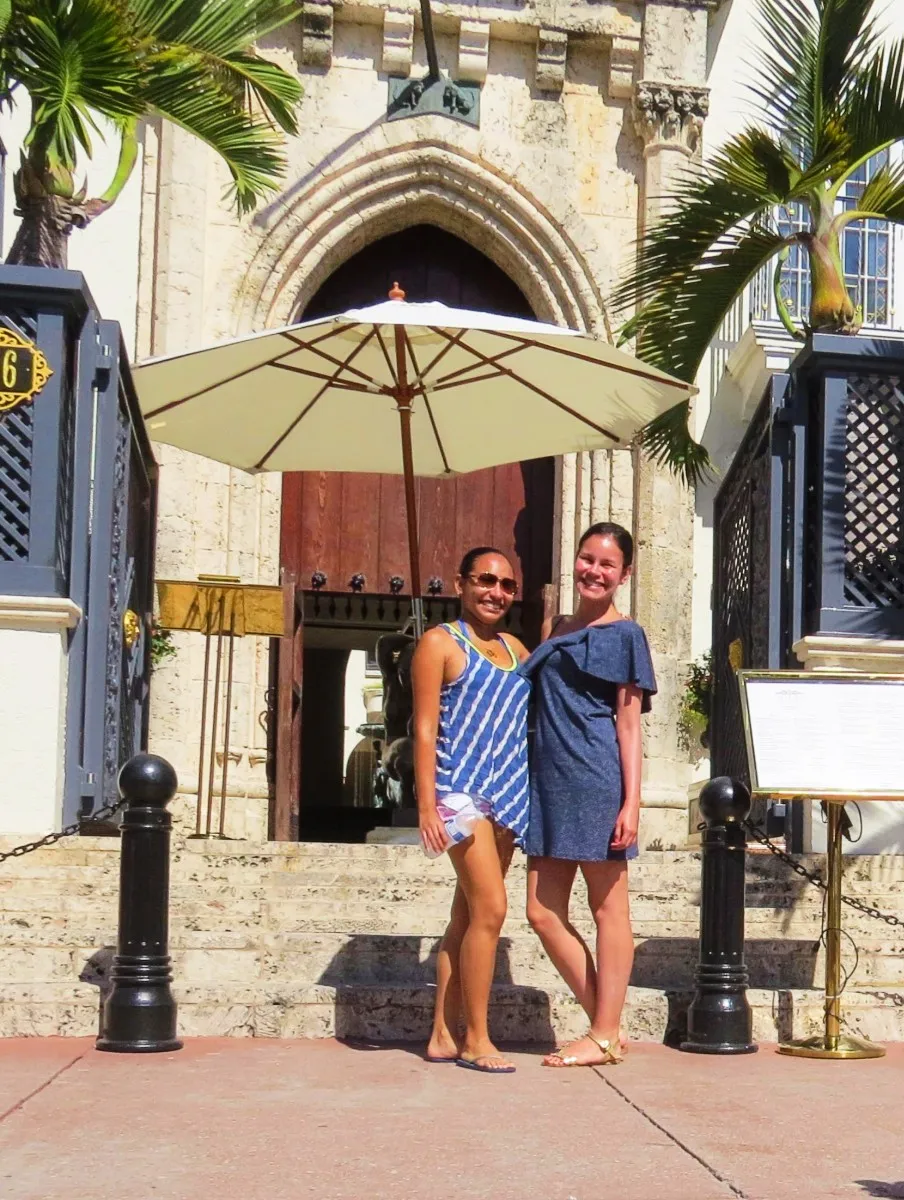 Two women smiling and posing under a large umbrella in front of a building with arched entrance and stone steps.