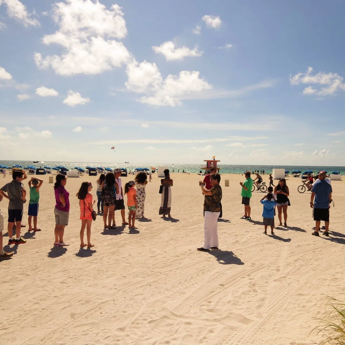Group of people standing and walking on a sunny sandy beach with umbrellas, a lifeguard tower, and the ocean in the background.