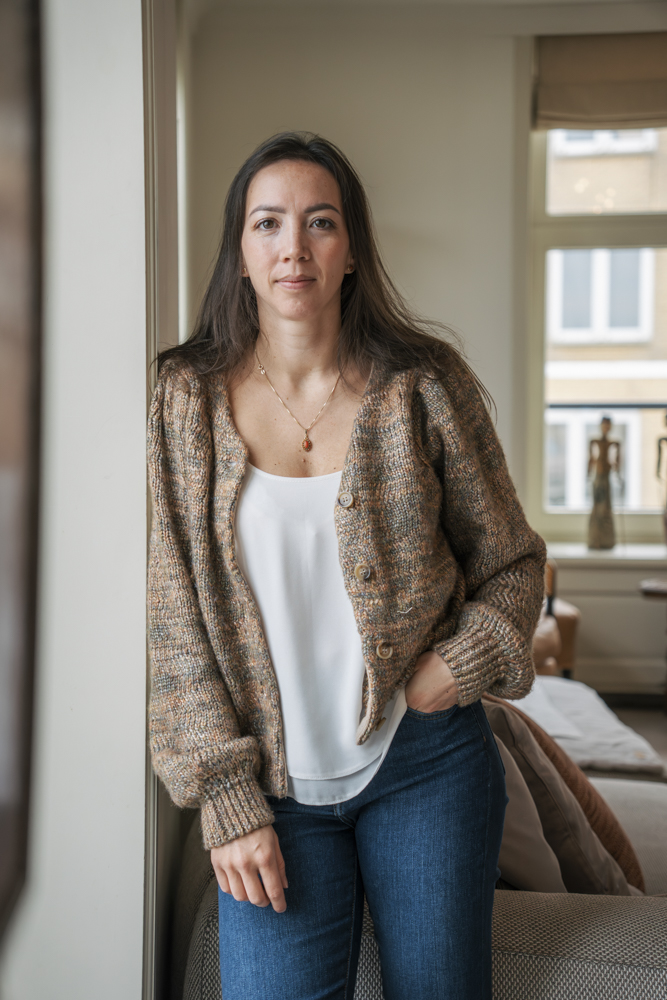 Vrouw met lang donker haar, een beige gebreid vest, witte top en blauwe jeans, staand bij een raam in een lichte woonkamer.