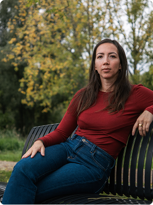 Vrouw met lang donker haar in een rood shirt en blauwe jeans zittend op een zwart parkbankje met groene bomen op de achtergrond.
