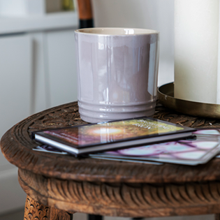 Grijze mok staat op een bruin, rond houten tafeltje naast een witte kaars en een stapel kleurrijke tijdschriften.