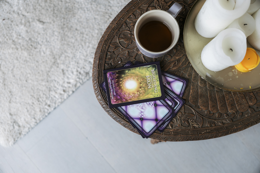 Houten tafel met een mok thee, stapel orakelkaarten en een schaal met witte en één gele kaars.
