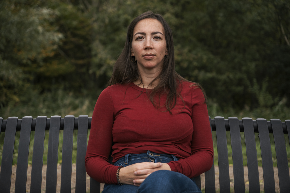 Vrouw in rood shirt en spijkerbroek zittend op een bankje in een groen park.