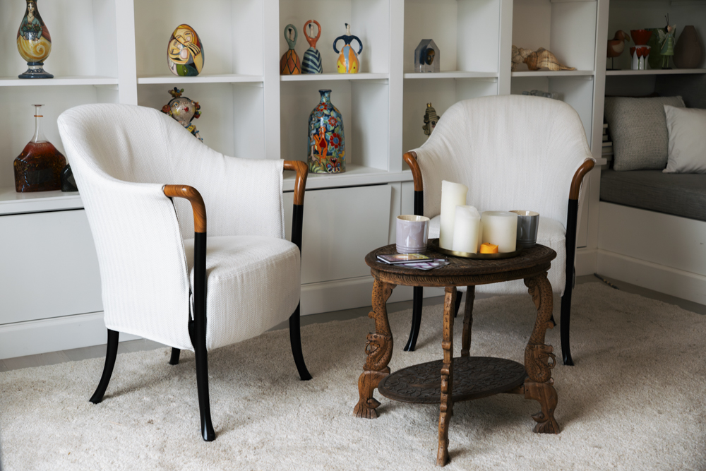 Twee witte stoelen met houten armleuningen rond een antiek houten tafeltje met kaarsen, in een lichte kamer met ingebouwde planken met kleurrijke keramische decoraties.