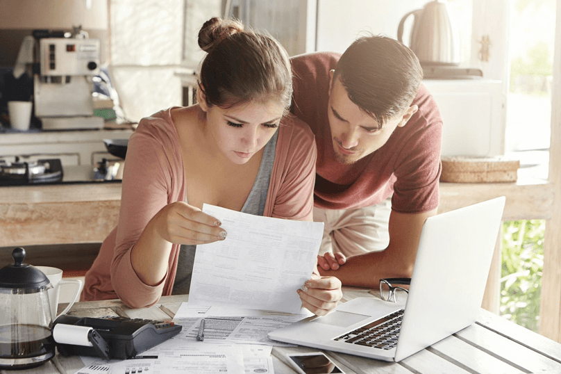 Una pareja joven sentada a una mesa revisando facturas y documentos financieros con una computadora portátil y una calculadora.