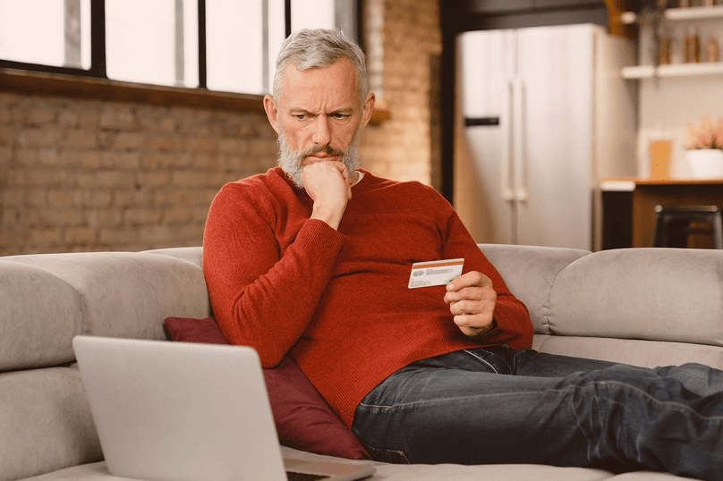 Hombre mayor sentado en un sillón mirando pensativamente su tarjeta de crédito mientras tiene una computadora portátil frente a él.