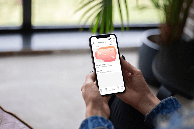 Persona sosteniendo un smartphone que muestra la sección de tarjetas virtuales de Banco del Sol en un entorno hogareño.