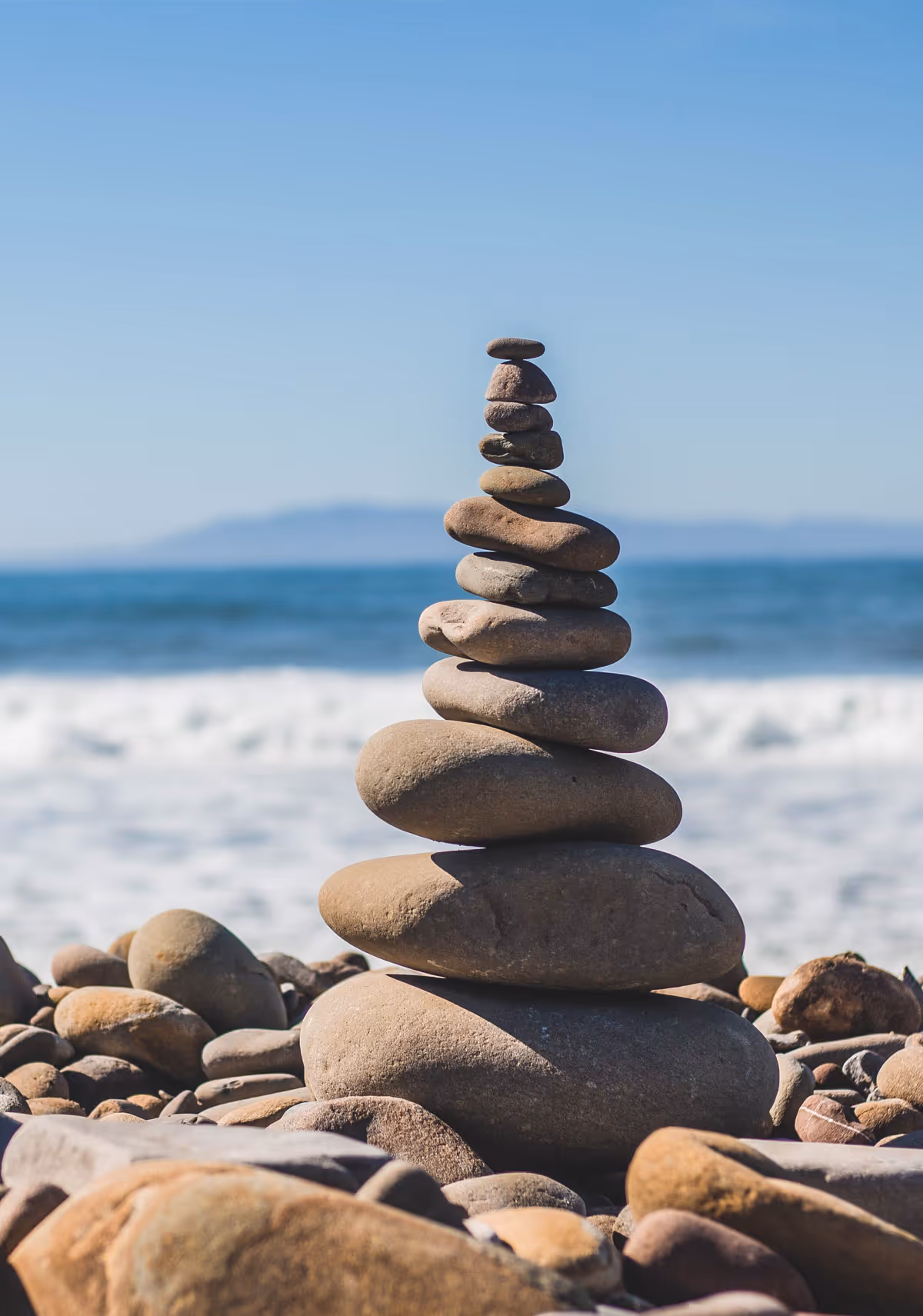 Stack of balanced smooth stones on a rocky beach with ocean waves and a clear blue sky in the background.