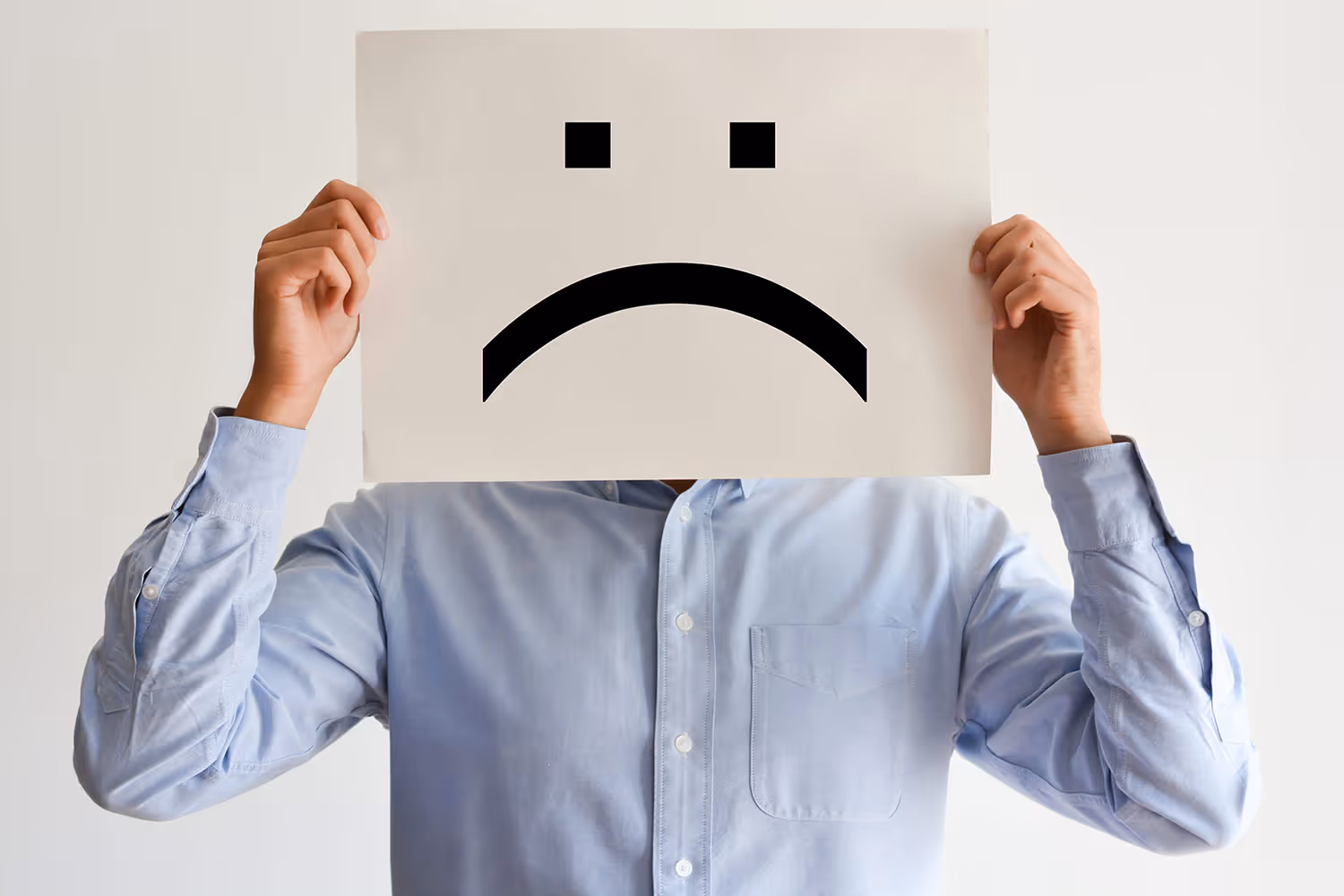 Person in light blue shirt holding a paper with a large black sad face emoji covering their own face.