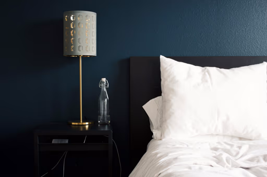 Nightstand with a modern gold lamp and a glass water bottle next to a bed with white pillows and sheets against a dark blue wall.
