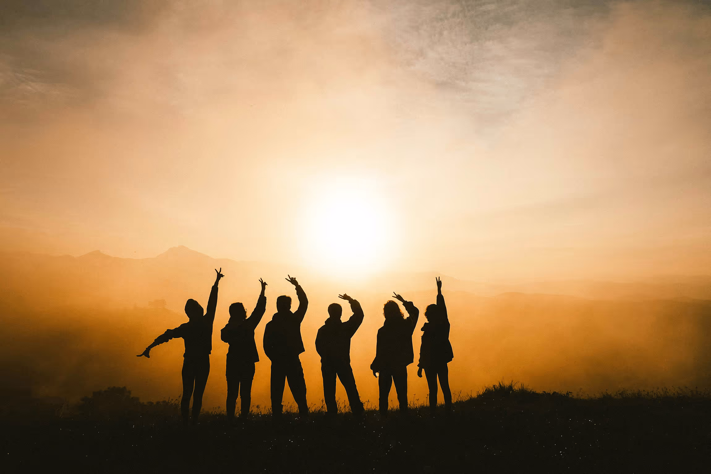 Silhouettes of six people standing on a hill with arms raised against a bright sunset sky.