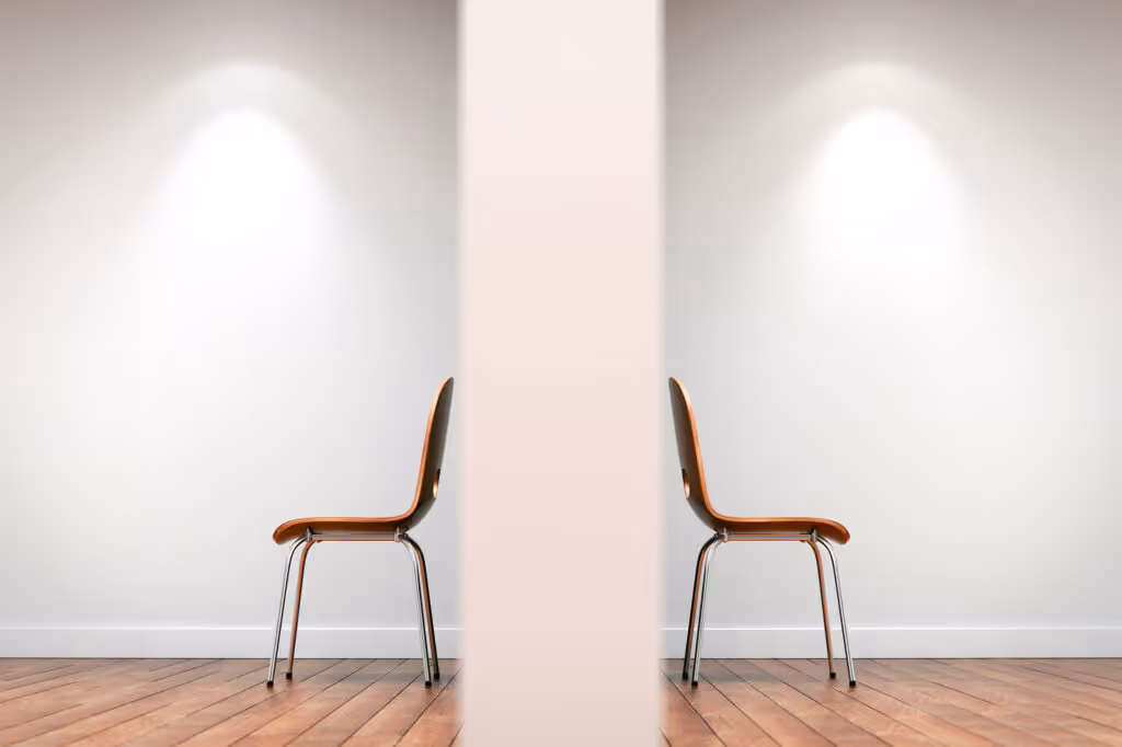 Two brown chairs facing away from each other separated by a central partition in a room with wooden floor and spotlighted white walls.