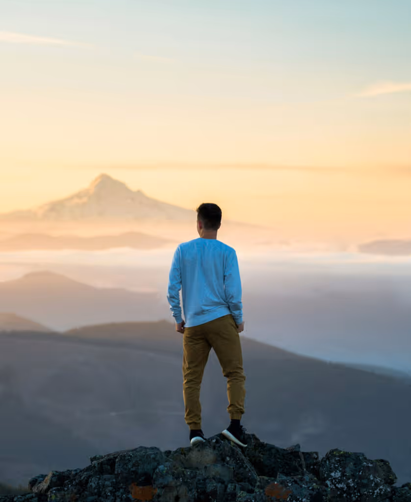 Man in light blue sweatshirt and tan pants standing on rocky mountain peak, looking at distant mountain during sunrise.