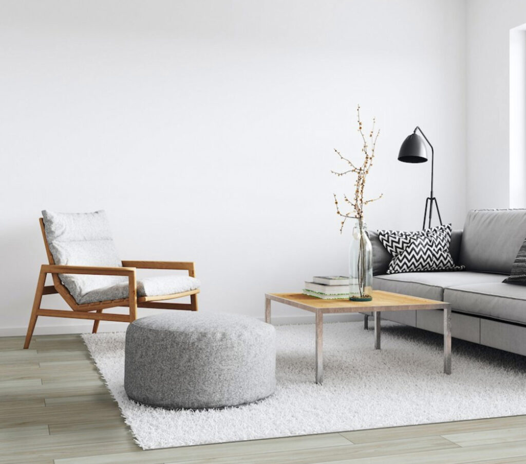 Modern living room with a wooden armchair, grey sectional sofa, round grey ottoman, light wooden coffee table with books and a vase, and a black floor lamp.