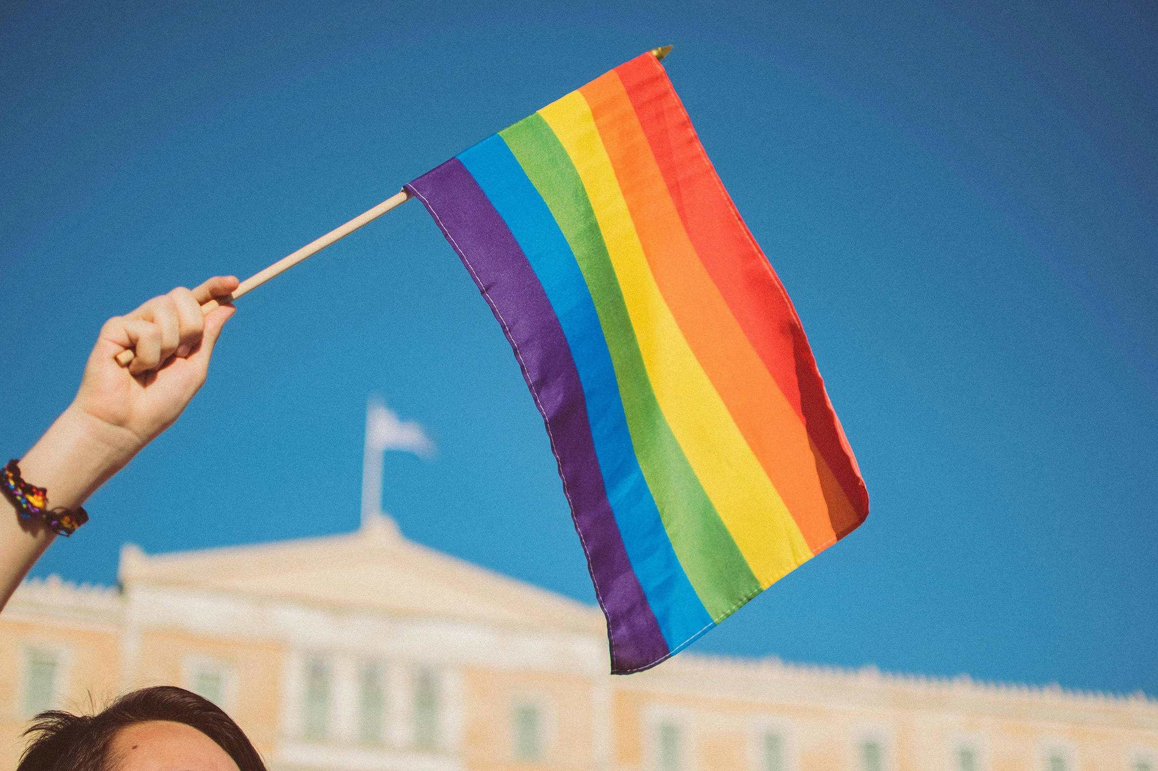 Hand holding a rainbow LGBTQ pride flag against a clear blue sky with a blurred building in the background.