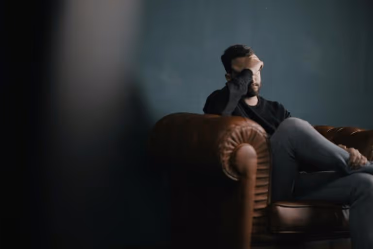 Man sitting on a brown leather armchair with his hand on his forehead looking distressed against a dark background.