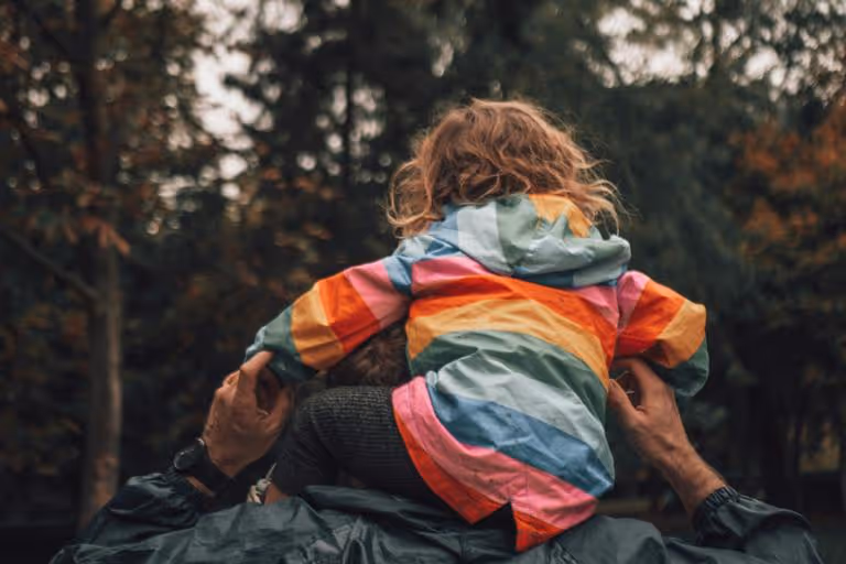 Child wearing a colorful striped jacket sitting on an adult's shoulders outdoors.
