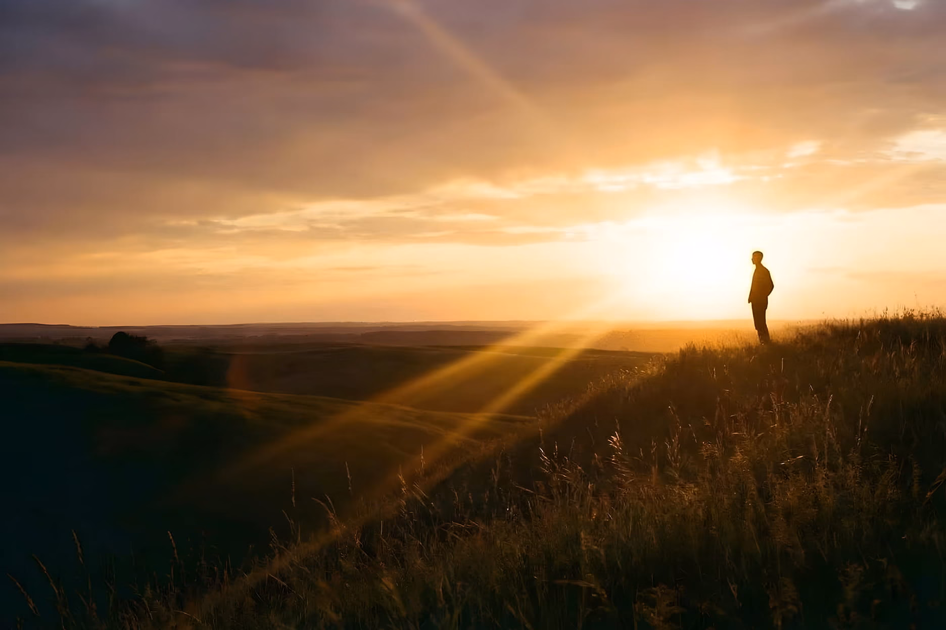 Person standing on a grassy hill overlooking a landscape at sunset with sun rays shining.