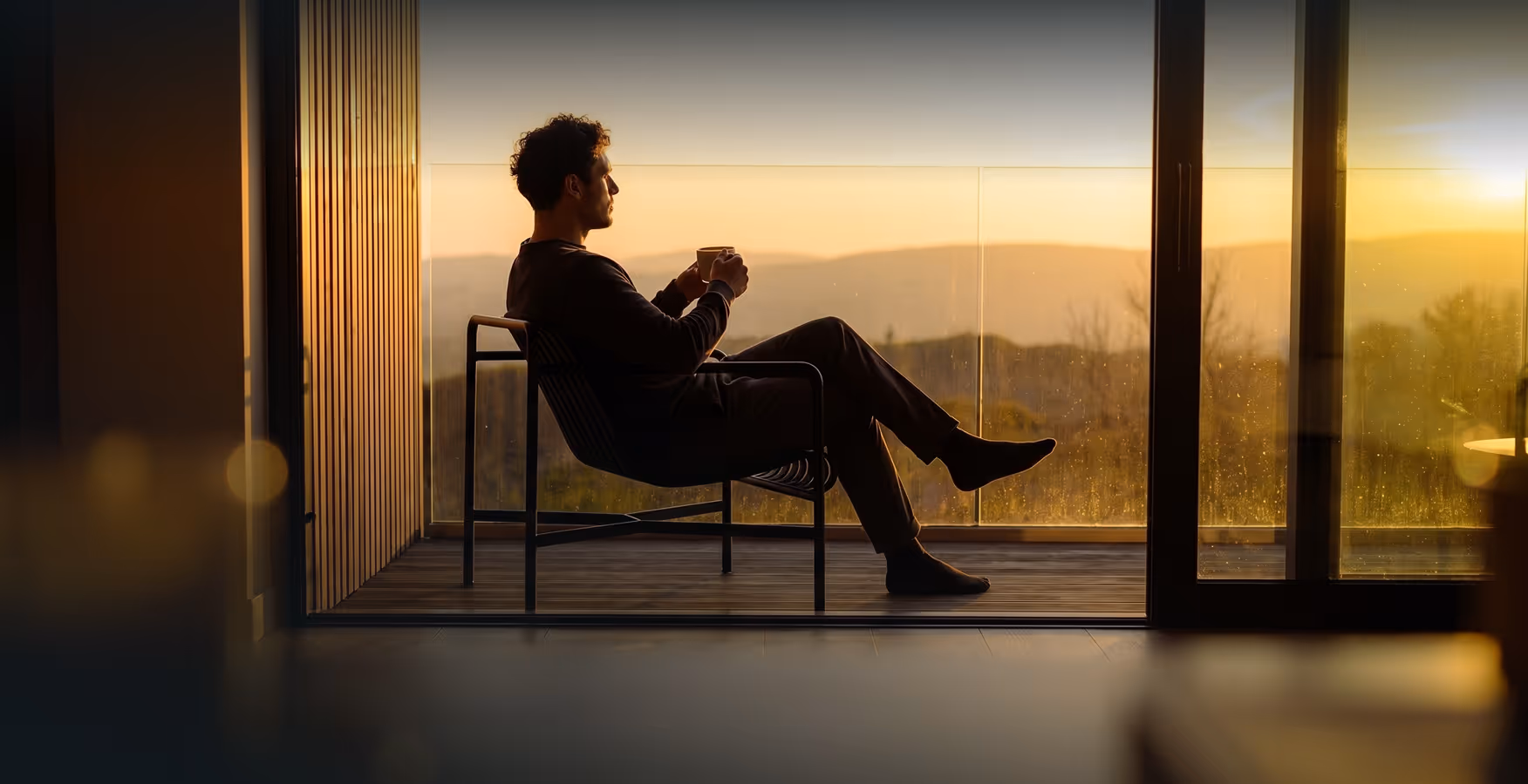 A man sitting on a chair on a balcony, holding a cup and looking out at a sunset over distant hills.