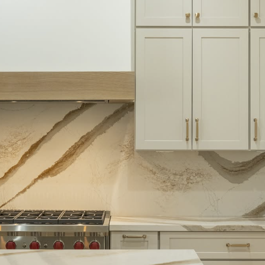 Modern kitchen countertop with beige and brown marble backsplash, stovetop with red knobs, and white cabinets with gold handles.