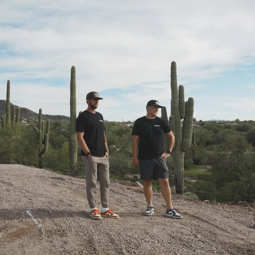 Two men wearing black t-shirts and caps standing on a dirt path with tall cacti and desert vegetation in the background under a cloudy sky.