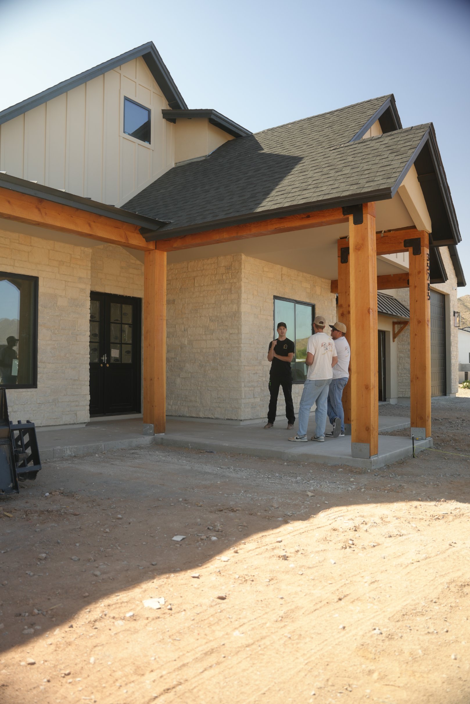 Three men standing and talking under the covered porch of a house with stone walls and wooden beams.