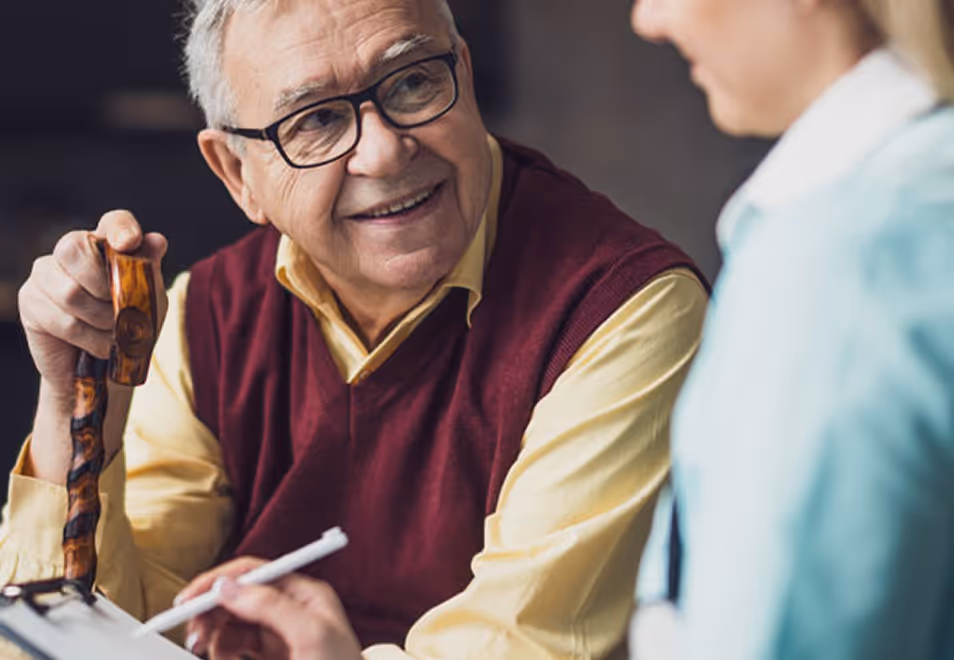Smiling elderly man with glasses holding a wooden cane, talking to a healthcare professional writing on a clipboard.