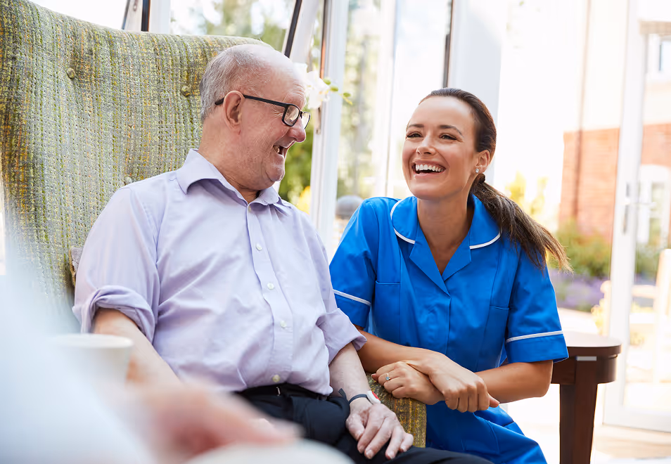 Smiling elderly man sitting in a green armchair talking with a cheerful female nurse in blue uniform in a bright room.