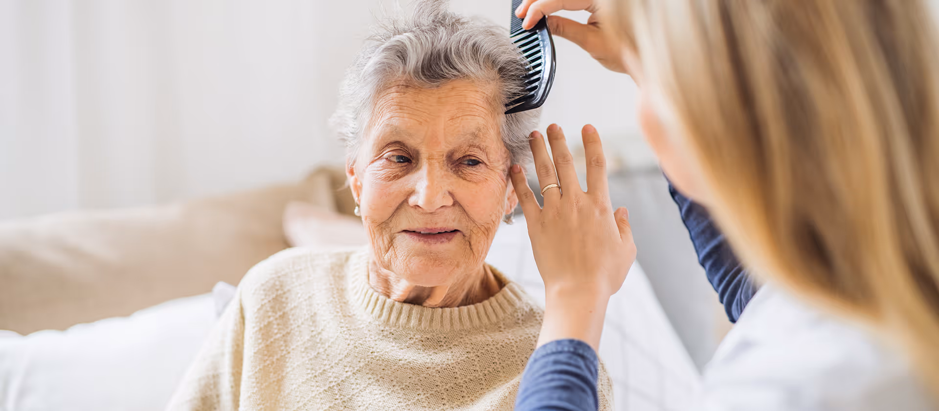 A woman combing the gray hair of an elderly woman wearing a beige sweater.