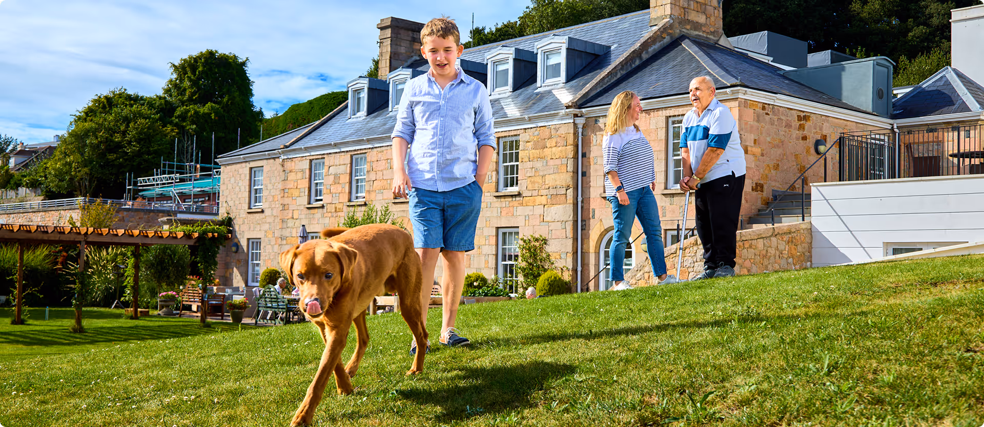A boy walks with a dog on a grassy hill in front of a stone building, while an older man and woman chat nearby.