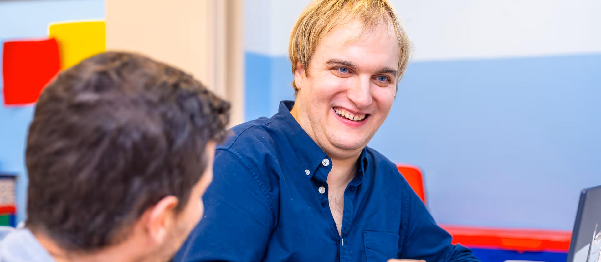 Smiling man with blonde hair in a blue shirt interacting with another person in a colorful room.