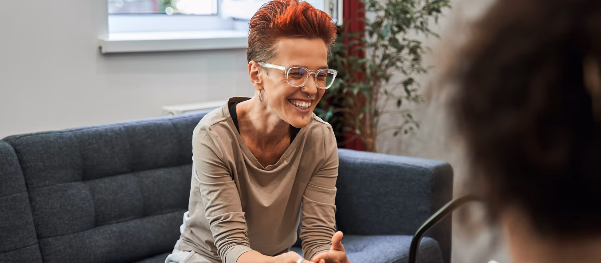 Smiling counselor with short red hair and glasses sitting on a couch, talking to a client.