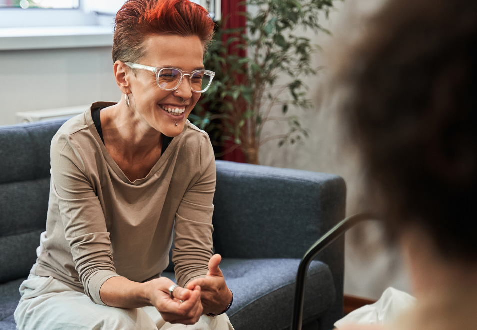 Smiling woman with short red hair and glasses talking to another person in a living room.