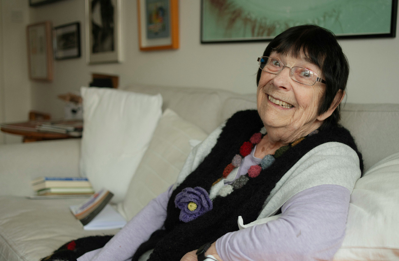 Smiling elderly woman with glasses sitting on a beige couch in a cozy living room.