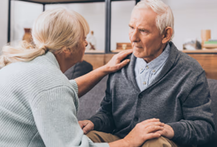 Concerned elderly woman comforting a distressed elderly man sitting on a couch in a home setting.