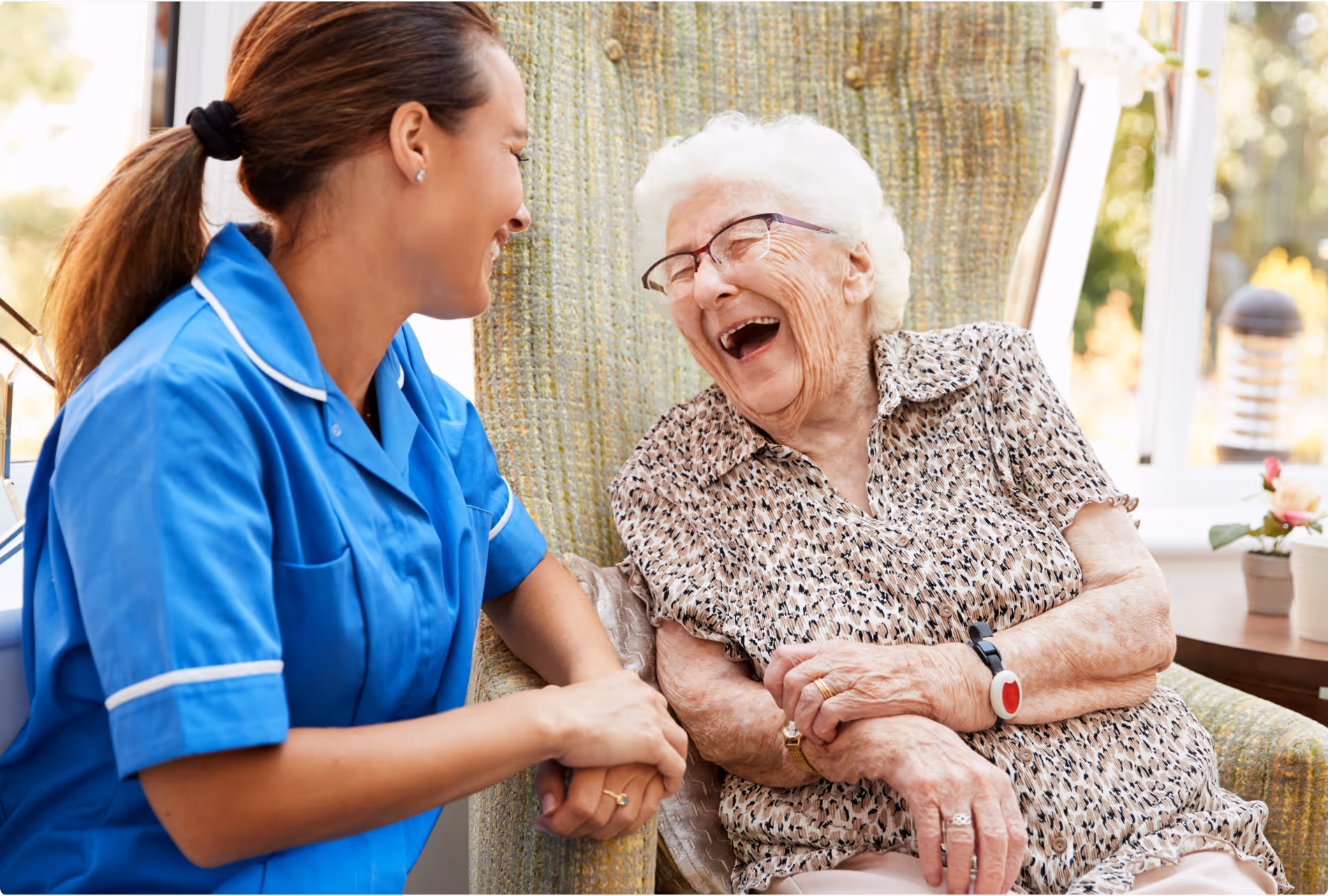 Caregiver in blue uniform holding hands and smiling with a laughing elderly woman in a patterned blouse seated in a chair.