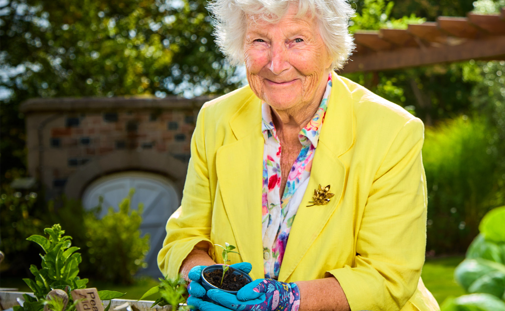 Smiling elderly woman in a yellow jacket and floral shirt holding a small potted plant while gardening outdoors.