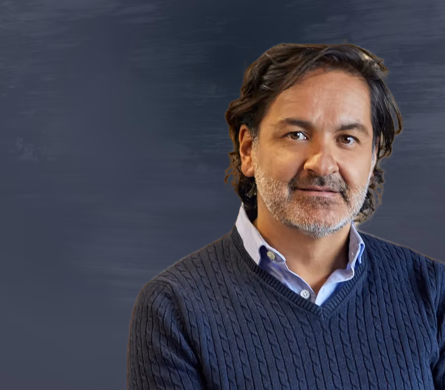 Middle-aged man with dark wavy hair and grey stubble wearing a blue sweater and light blue collared shirt, standing against a dark grey background.