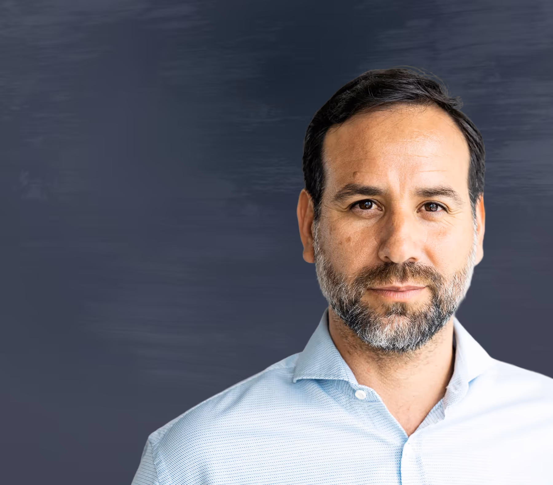Confident man with short dark hair and beard wearing a light blue shirt against a dark background.
